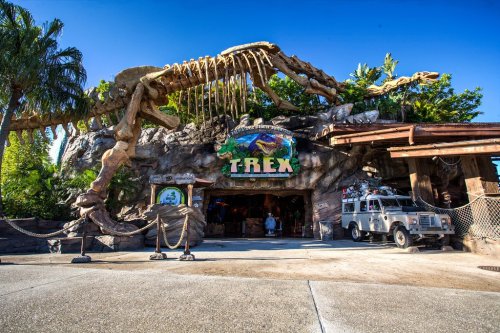 Exterior of T-Rex restaurant featuring a dinosaur skeleton and a rocky entrance, with palm trees and a clear blue sky.