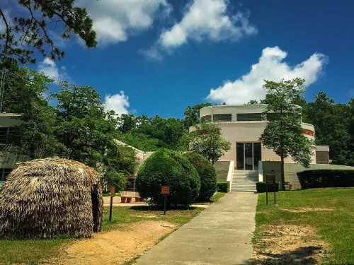 A modern building surrounded by greenery and a thatched hut under a blue sky with fluffy clouds.