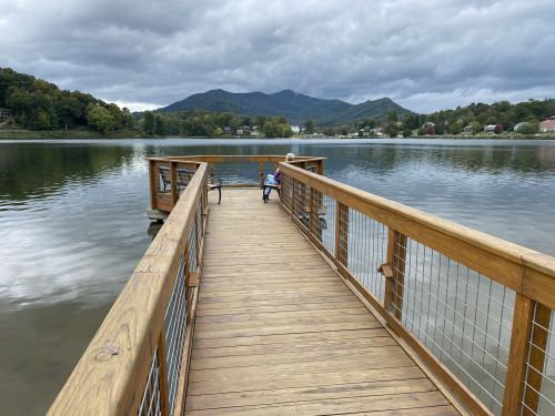 A wooden dock extends over calm water, with a child sitting at the end, surrounded by mountains and cloudy skies.