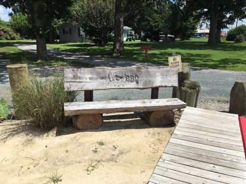 A rustic wooden bench with a BBQ logo, set in a grassy area near a pathway and trees.