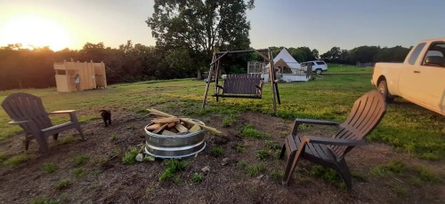 A peaceful outdoor scene with a fire pit, wooden chairs, a swing, and a truck, set against a sunset backdrop.