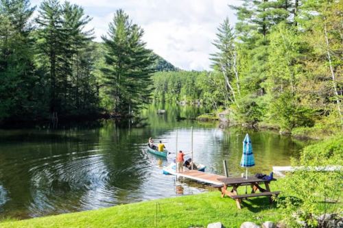 A serene lake scene with a dock, a canoe, and people enjoying nature surrounded by lush green trees.