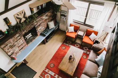 Cozy living room with a brick fireplace, orange chairs, a wooden coffee table, and a patterned rug. Bright and inviting space.