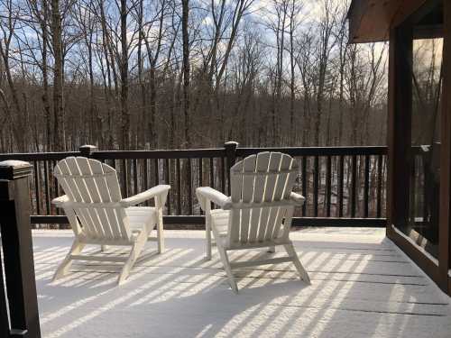 Two white Adirondack chairs on a snowy deck, overlooking a forest of bare trees under a clear sky.