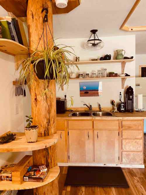 Cozy kitchen with wooden shelves, a large support post, plants, and a view of a scenic photo on the wall.