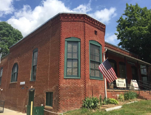 Historic red brick building with green shutters, steps, and an American flag in front, surrounded by greenery.