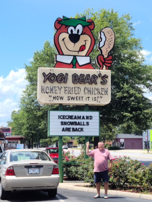 A man stands next to a sign for Yogi Bear's Honey Fried Chicken, featuring a cartoon bear and announcing ice cream and snowballs.