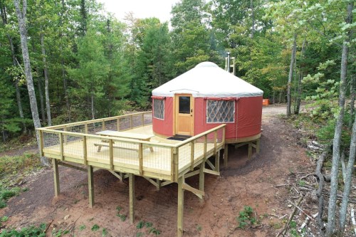 A red yurt on a wooden deck surrounded by trees in a forested area.