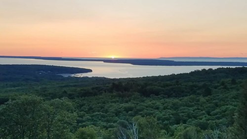 A serene landscape at sunset, featuring a lake surrounded by lush greenery and distant hills under a colorful sky.
