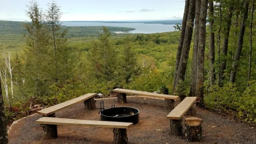 A scenic view of a fire pit surrounded by wooden benches, overlooking a lush green landscape and a lake in the distance.