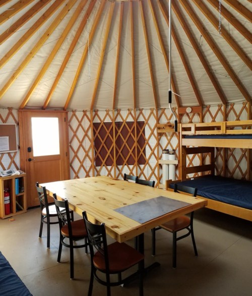 Interior of a yurt featuring wooden beams, a table with chairs, and bunk beds against the walls.
