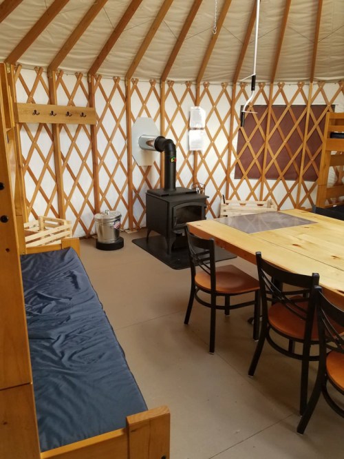 Interior of a yurt featuring a wooden table, chairs, a wood stove, and a cozy seating area.