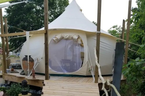A white tent with a pointed top, set on a wooden deck surrounded by greenery and decorative elements.