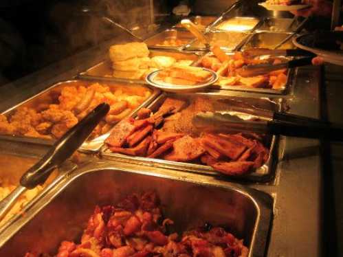 A buffet display featuring various cooked dishes, including fried items and sides, with steam rising from the trays.