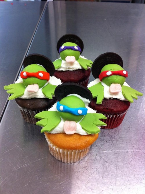 Five colorful cupcakes topped with frog figures wearing sunglasses and hats, each with a unique frosting color.