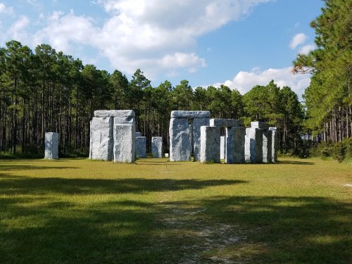 A stone structure resembling Stonehenge, surrounded by trees and grass under a partly cloudy sky.
