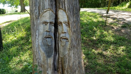 Carved tree trunk featuring two faces with exaggerated expressions, set in a green, grassy landscape.