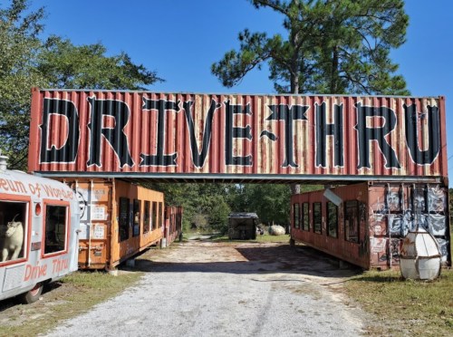 A weathered drive-thru entrance with large letters, surrounded by vintage vehicles and trees under a clear blue sky.
