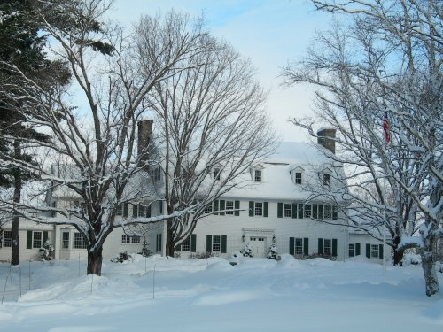 A large white house surrounded by snow-covered trees on a winter day, with a clear blue sky in the background.