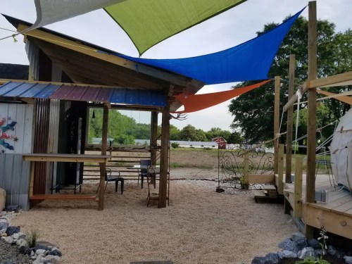 A cozy outdoor space with colorful shade sails, gravel flooring, and wooden structures, surrounded by greenery.