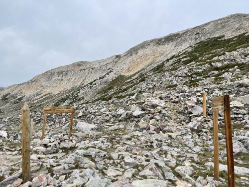 Rocky landscape with wooden posts marking a trail, surrounded by steep hills under a cloudy sky.