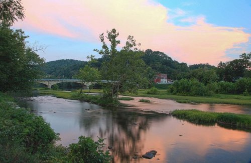 A serene river scene at sunset, featuring a bridge, lush greenery, and soft reflections on the water.
