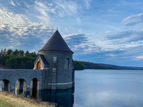 A stone tower with a conical roof stands by a calm lake, surrounded by trees and a cloudy sky.
