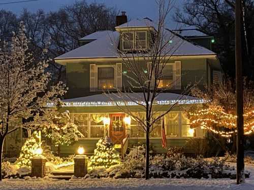 A cozy green house adorned with snow, holiday lights, and decorations, surrounded by a winter landscape at dusk.