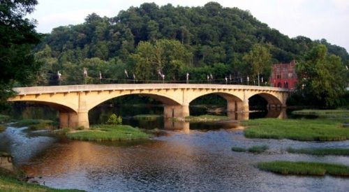 A stone bridge spans a calm river, surrounded by lush greenery and a hillside in the background.