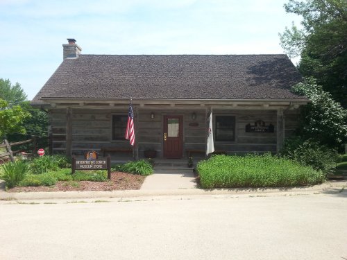 A rustic log cabin with a red door, American flag, and signs for a museum, surrounded by greenery and a clear sky.