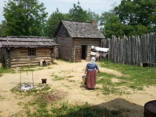 A woman in period clothing walks towards a rustic log cabin in a fenced yard with trees in the background.