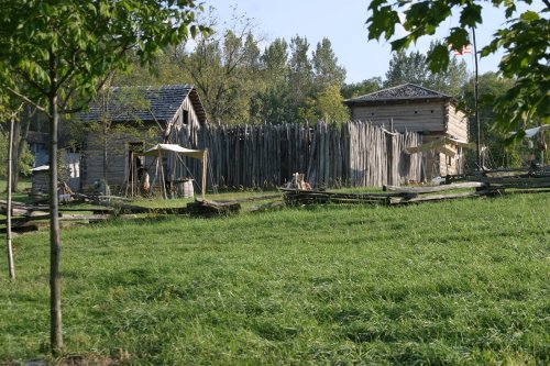 A historic settlement with wooden buildings and a palisade, surrounded by lush green grass and trees.