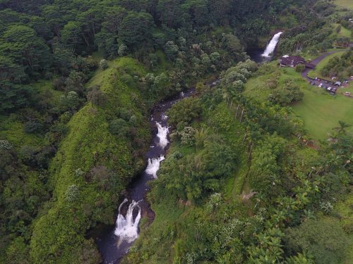 Aerial view of lush green hills with a winding river and waterfalls, surrounded by dense vegetation.