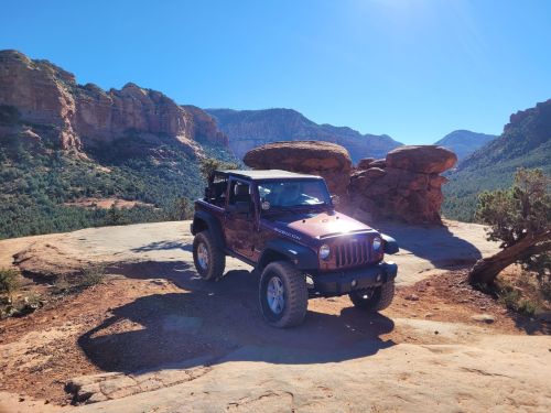 A purple Jeep parked on a rocky outcrop with scenic mountains and clear blue skies in the background.