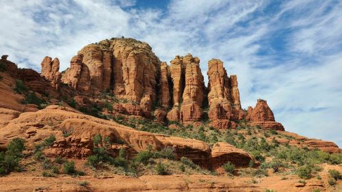 Red rock formations rise against a blue sky with scattered clouds, showcasing the rugged beauty of the landscape.