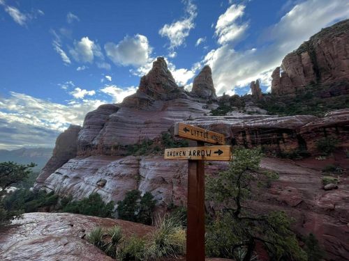 A wooden signpost with directions to "Little Horse" and "Broken Arrow" in a rocky landscape under a cloudy sky.