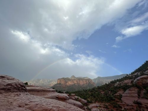 A scenic view of a rainbow arching over rocky mountains under a partly cloudy sky.