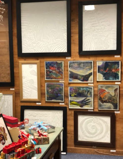 A gallery wall featuring various framed artworks, some with abstract designs, alongside a table with gift items.
