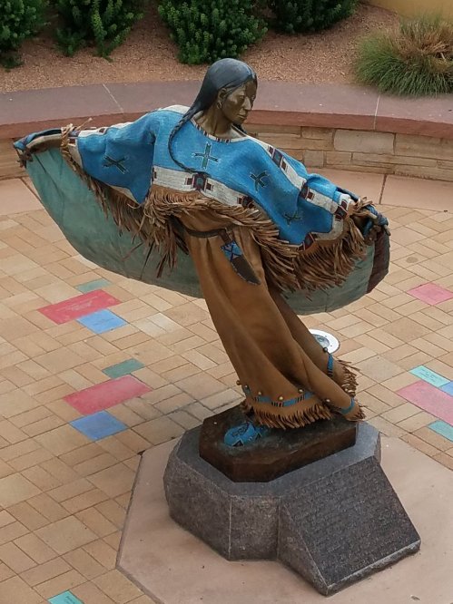 A bronze statue of a Native American woman in traditional attire, gracefully holding a flowing shawl.