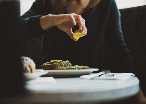A person squeezes lemon over a plate of avocado toast at a café table.