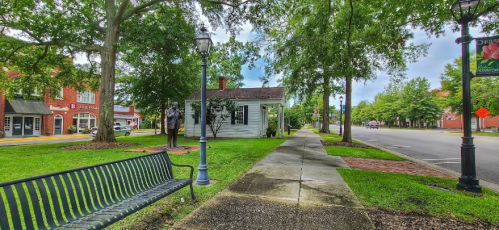A tree-lined street with a bench, a small white house, and shops in the background under a partly cloudy sky.