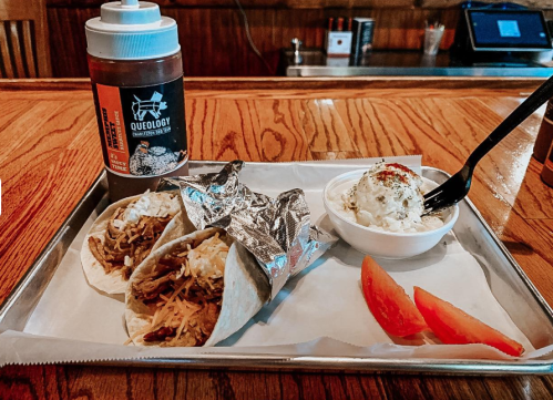 Two tacos with shredded meat and cheese, a side of ice cream, and a bottle of sauce on a wooden table.