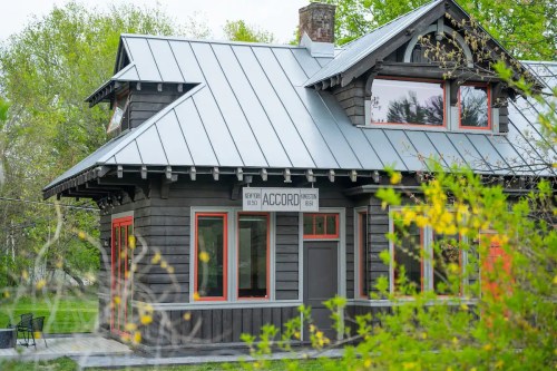 A charming dark wooden house with a metal roof and red accents, featuring a sign that reads "Accord." Green foliage surrounds it.