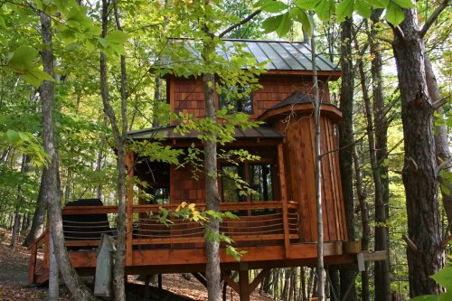 A charming wooden treehouse nestled among green trees, featuring a sloped roof and a wraparound deck.