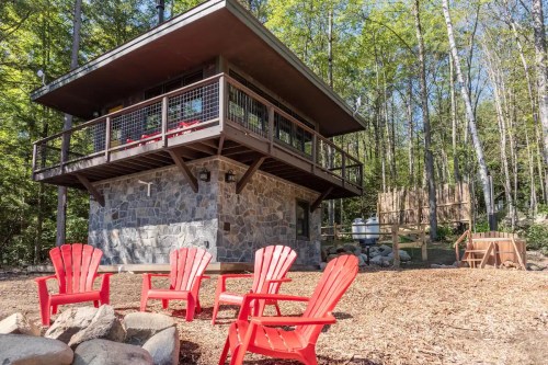 A modern cabin with a stone base surrounded by trees, featuring red Adirondack chairs and a fire pit in front.