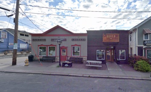 A street view of a bakery with a red and gray facade, benches outside, and a cloudy sky in the background.