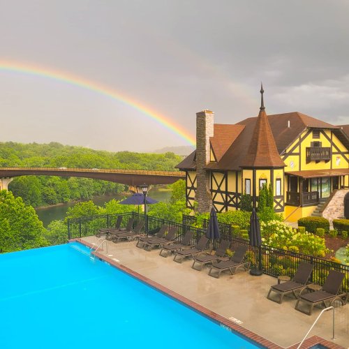 A scenic view of a poolside with a rainbow over a river and a charming house in the background.
