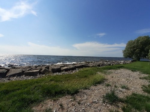 A serene lakeside view with rocky shore, green grass, and a clear blue sky with scattered clouds.