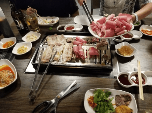 A table set for a barbecue meal with various meats, sauces, and side dishes, with someone holding a plate of pink meat.