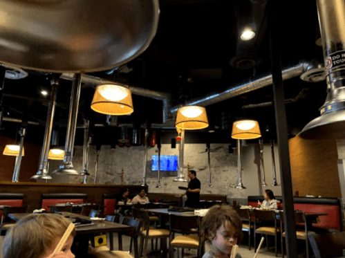 A modern restaurant interior with hanging lamps, metal accents, and diners at tables. A server is visible in the background.